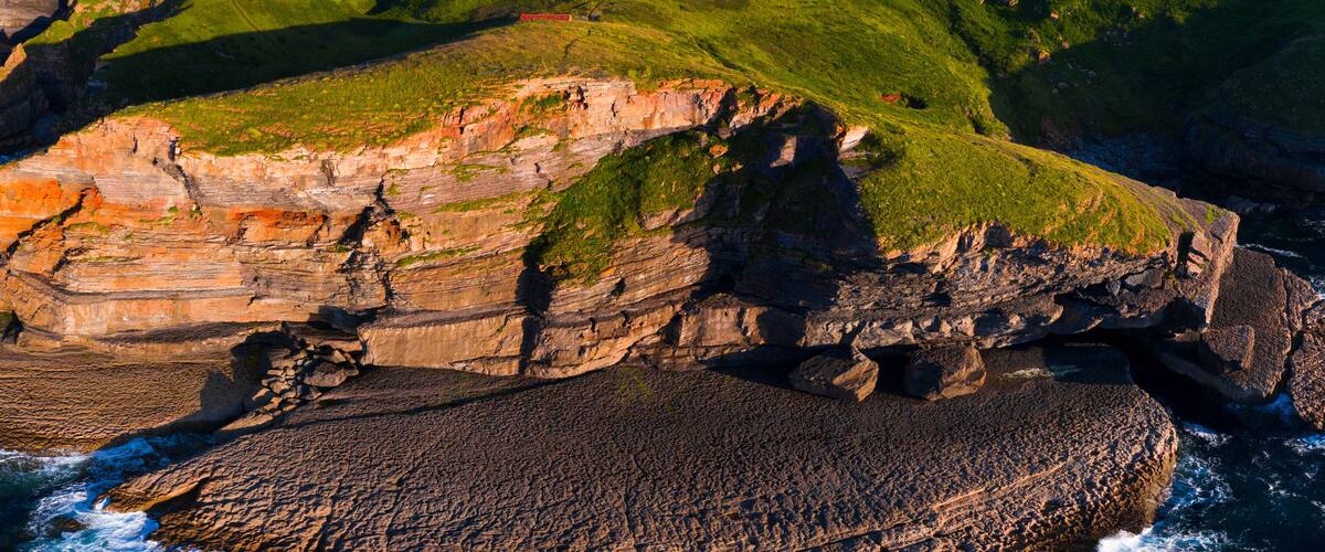 A coastal cliff with layered rock formations and a grassy hill in the background.