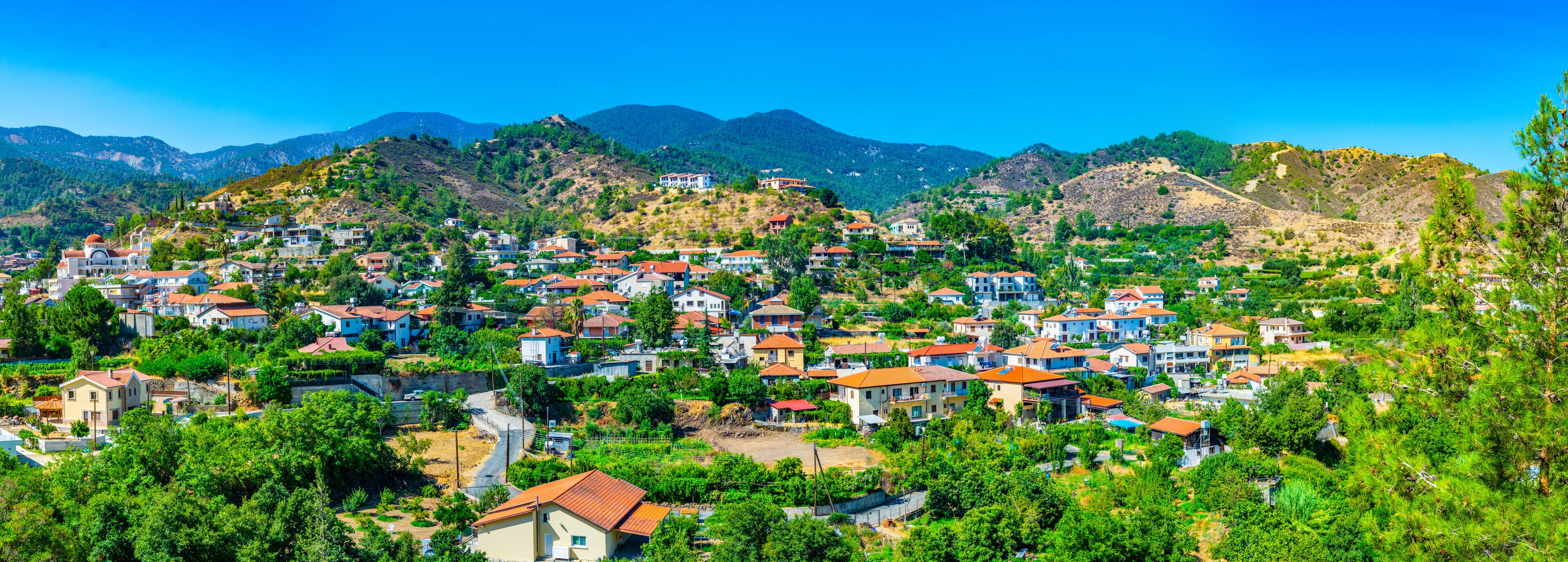 A hillside village with red-roofed houses, lush greenery, and mountains in the background.