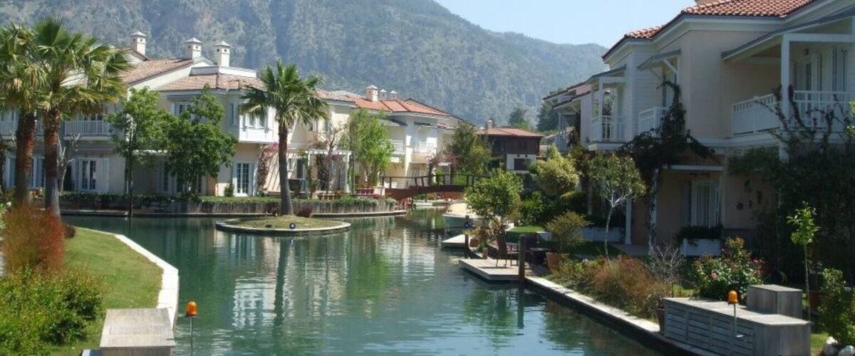 A canal with buildings and palm trees, surrounded by mountains.