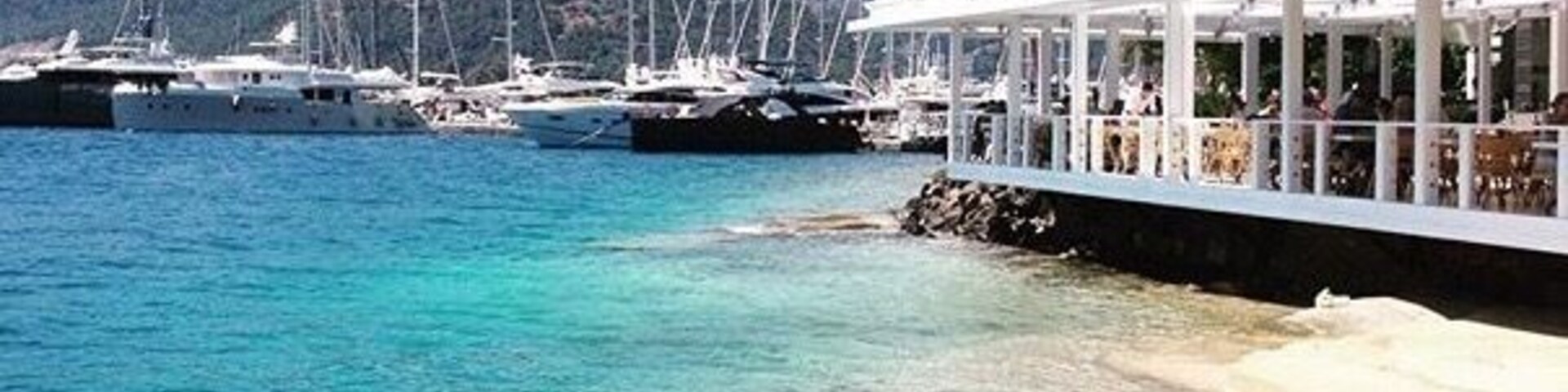 Un puerto deportivo con veleros amarrados, una playa de arena y montañas al fondo.