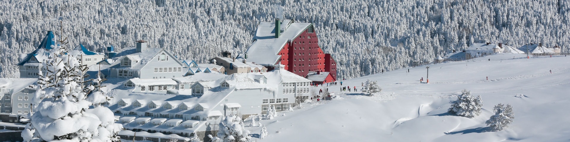 Un pueblo cubierto de nieve con un edificio destacado de techo rojo, un bosque denso y un cielo azul despejado.