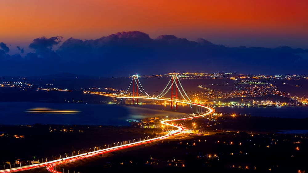 Un pont aux câbles illuminés, une route sinueuse et une vue sur la ville au crépuscule.