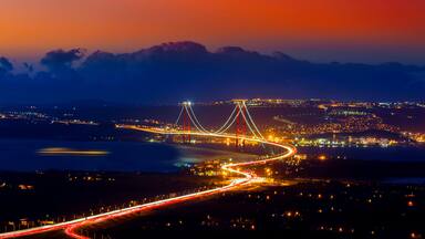 Un puente con cables iluminados, una carretera serpenteante y un paisaje urbano al anochecer.