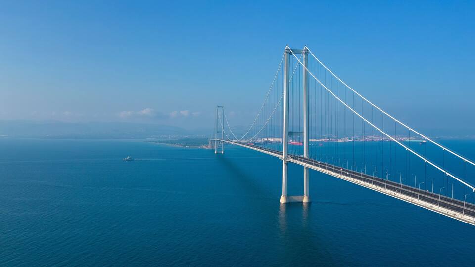 A large suspension bridge spanning a deep blue body of water, with a clear sky in the background.
