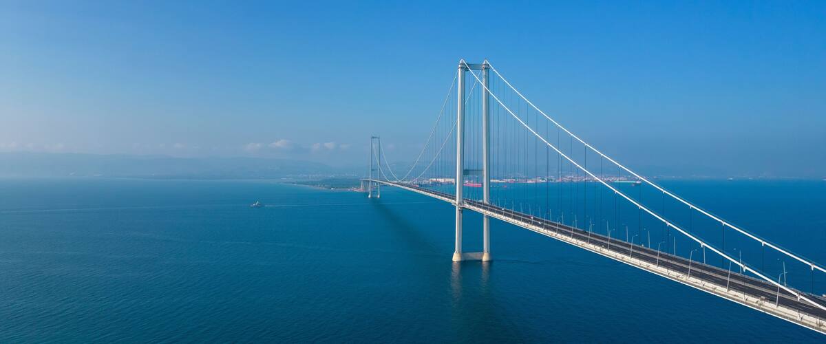A large suspension bridge spanning a deep blue body of water, with a clear sky in the background.