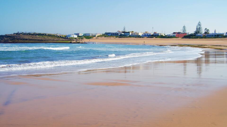 A beach with waves, a sandy shore, and buildings in the background.