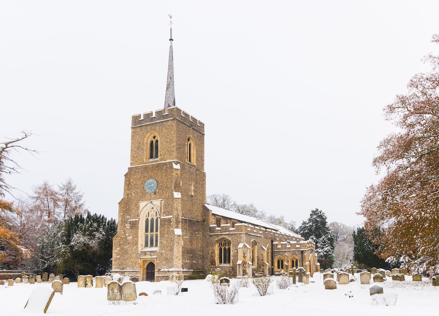 Una iglesia con una aguja alta y un reloj, rodeada de árboles cubiertos de nieve y lápidas.