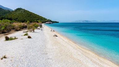 A pebble beach with clear turquoise water, a person walking, and a green hill in the background.