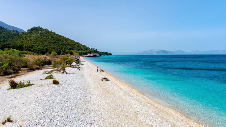 A pebble beach with clear turquoise water, a person walking, and a green hill in the background.