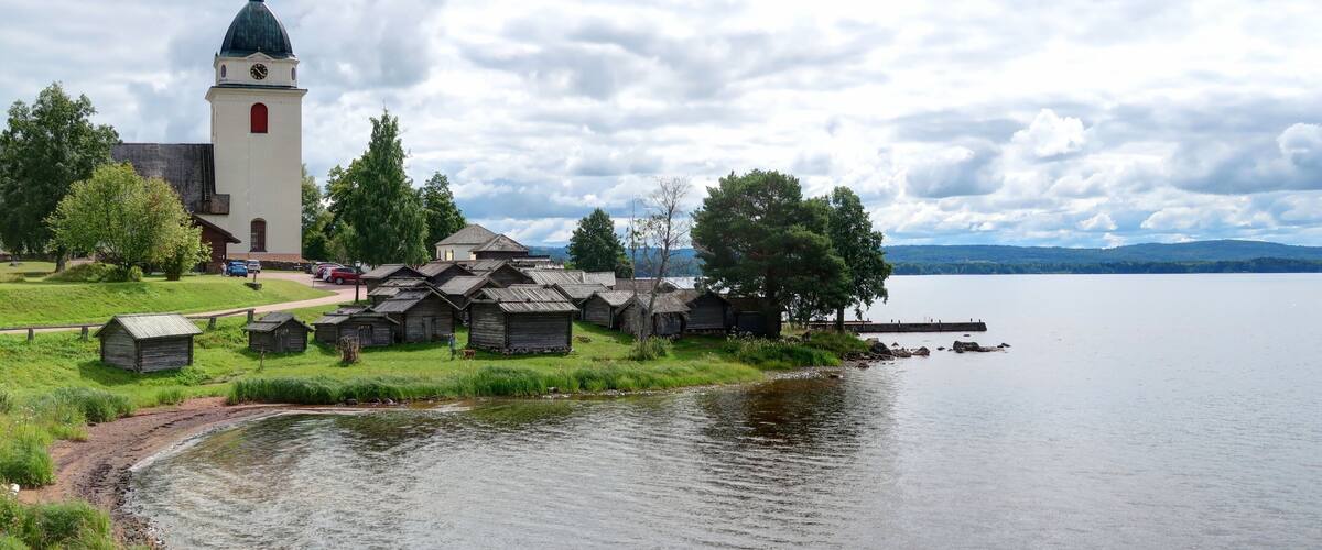 A church with a tower, wooden structures, a lake, and a forested area.