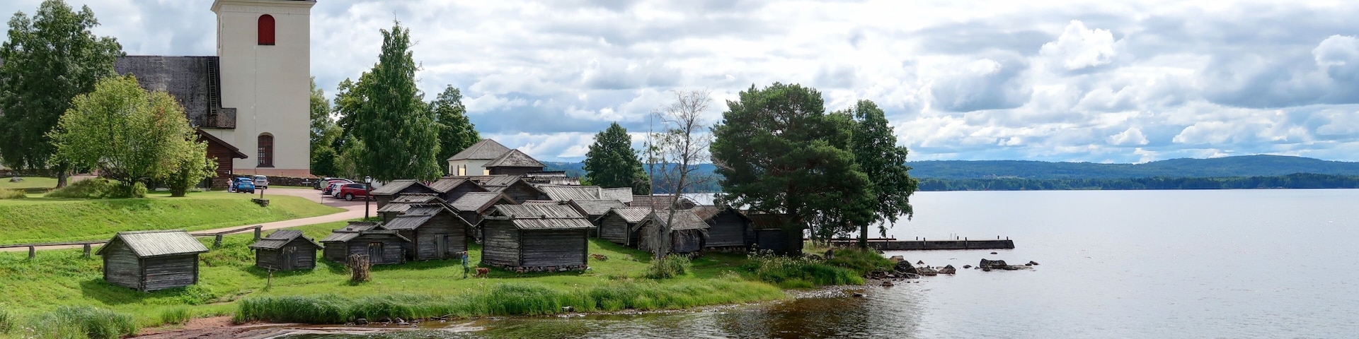 A church with a tower, wooden structures, a lake, and a forested area.
