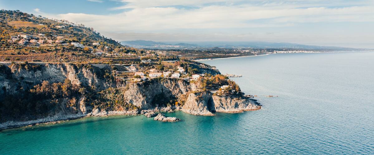 A coastal landscape with a rocky cliff, turquoise sea, and scattered houses.