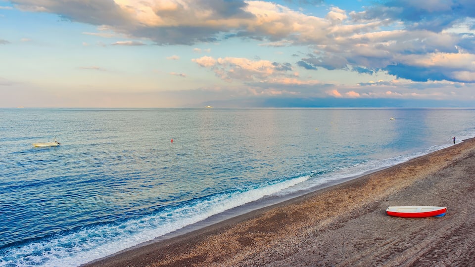 A beach with a boat on the shore, a distant ship, and a person fishing.