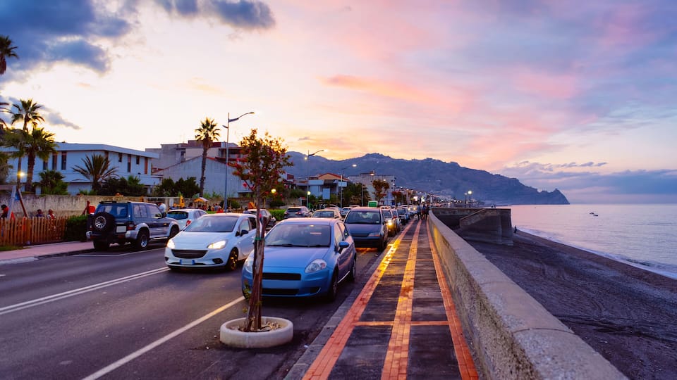 A coastal road with parked cars, palm trees, and a sunset over the sea.