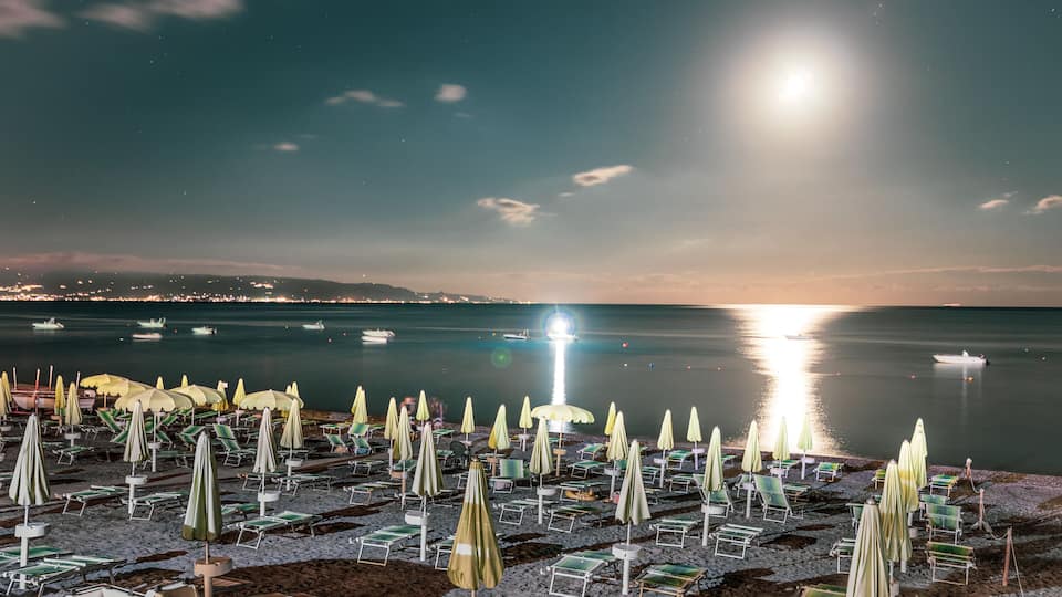 A beach at night with rows of sun loungers and umbrellas under a bright moon.