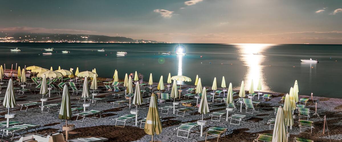 A beach at night with rows of sun loungers and umbrellas under a bright moon.