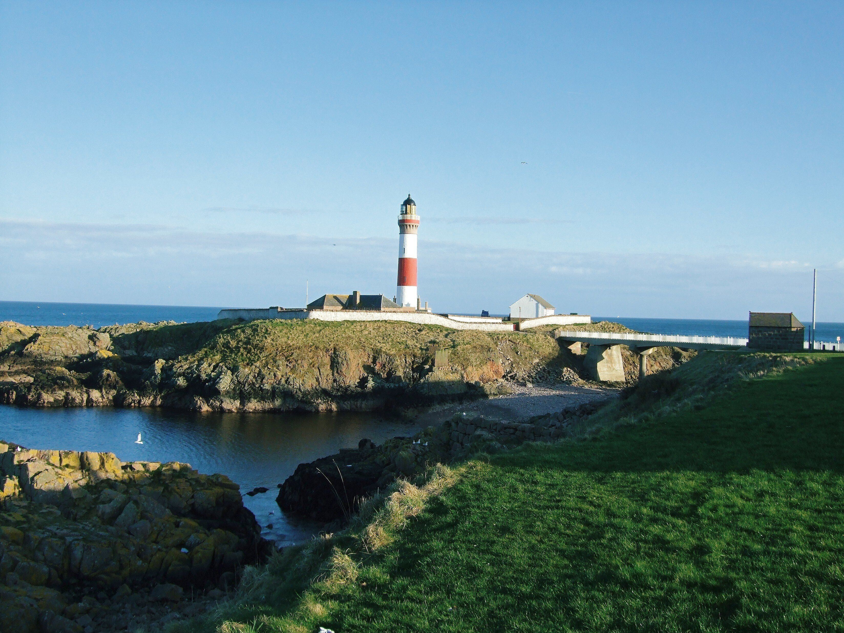 A lighthouse on a rocky outcrop with a bridge leading to a white building.