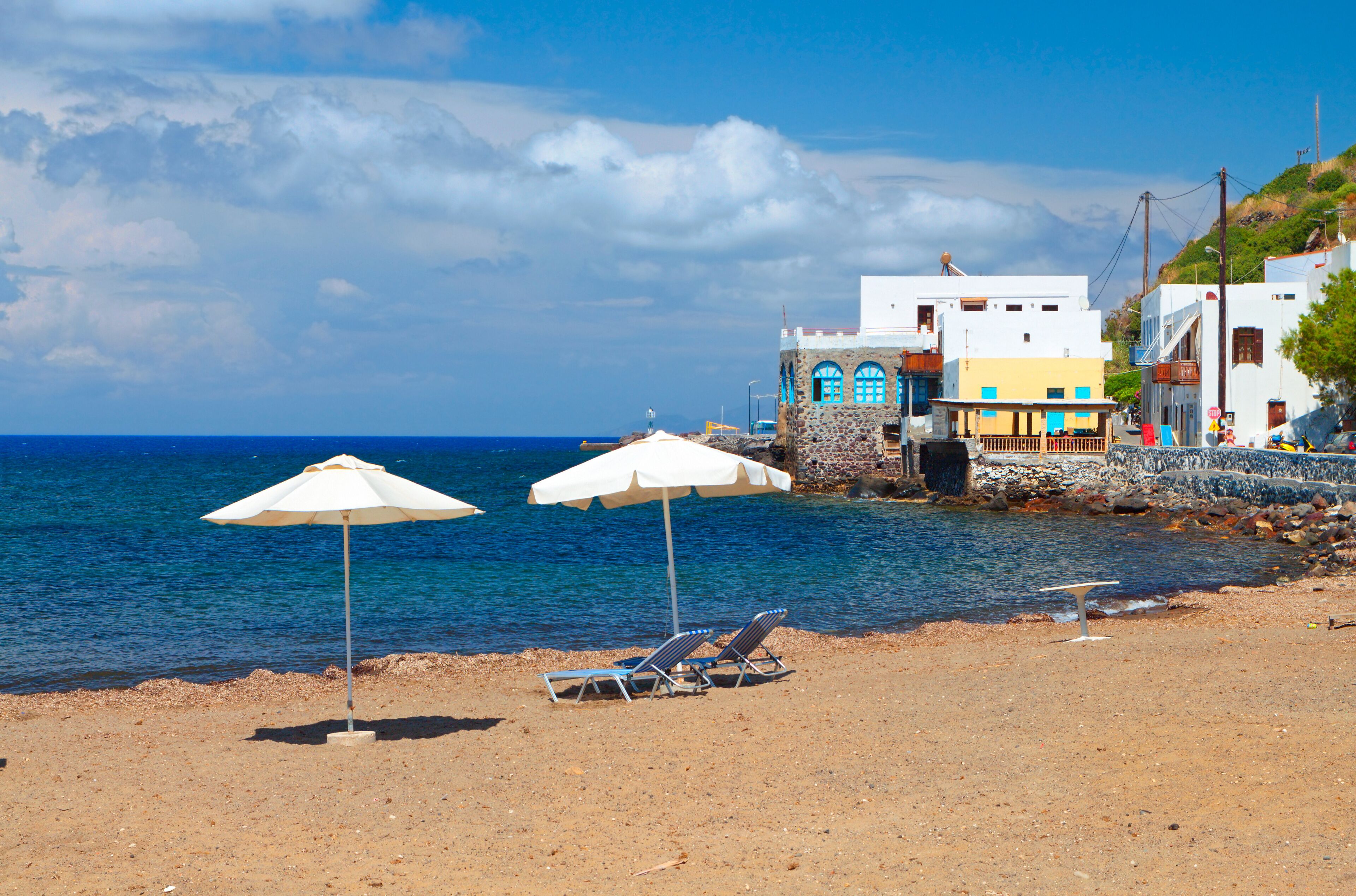Une plage avec deux parasols blancs, une côte rocheuse et, en arrière-plan, des bâtiments aux couleurs vives.