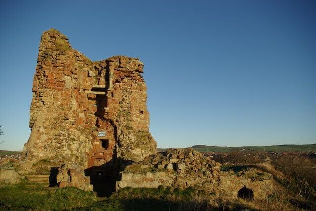 A large, ancient stone structure with a central tower and a surrounding wall, set in a grassy field with a clear sky.