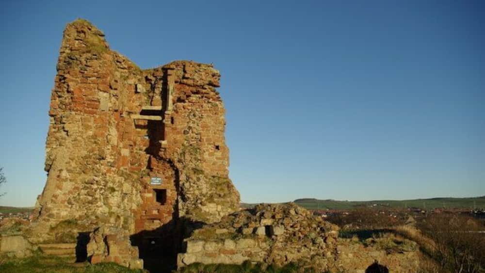 A large, ancient stone structure with a central tower and a surrounding wall, set in a grassy field with a clear sky.