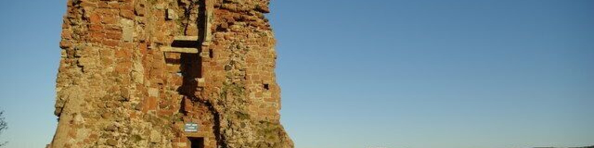 Una gran estructura de piedra antigua con una torre central y un muro que la rodea, ubicada en un campo cubierto de hierba bajo un cielo despejado.