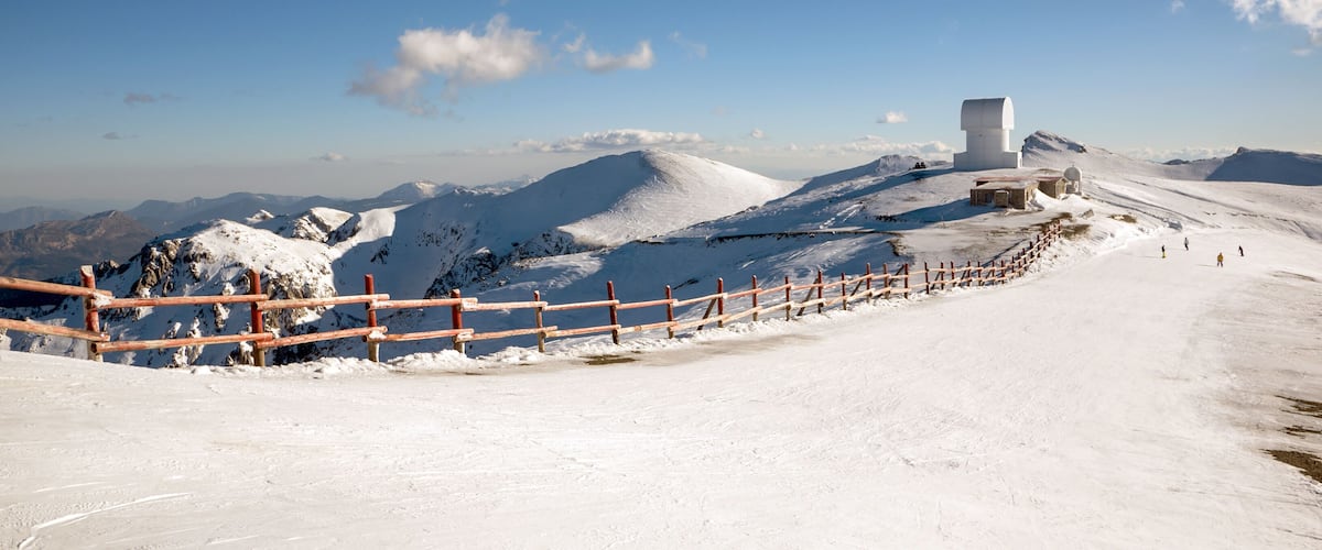 A snowy mountain landscape with a wooden fence, a white building, and distant mountains under a clear blue sky.
