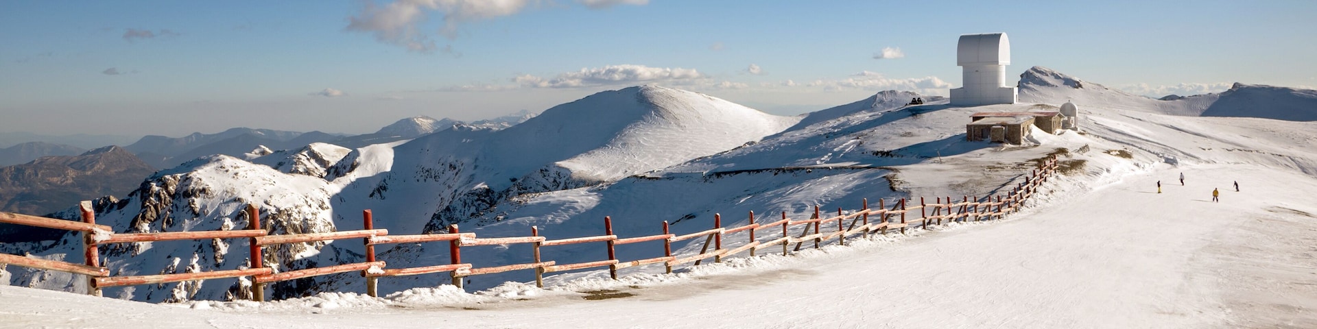 A snowy mountain landscape with a wooden fence, a white building, and distant mountains under a clear blue sky.