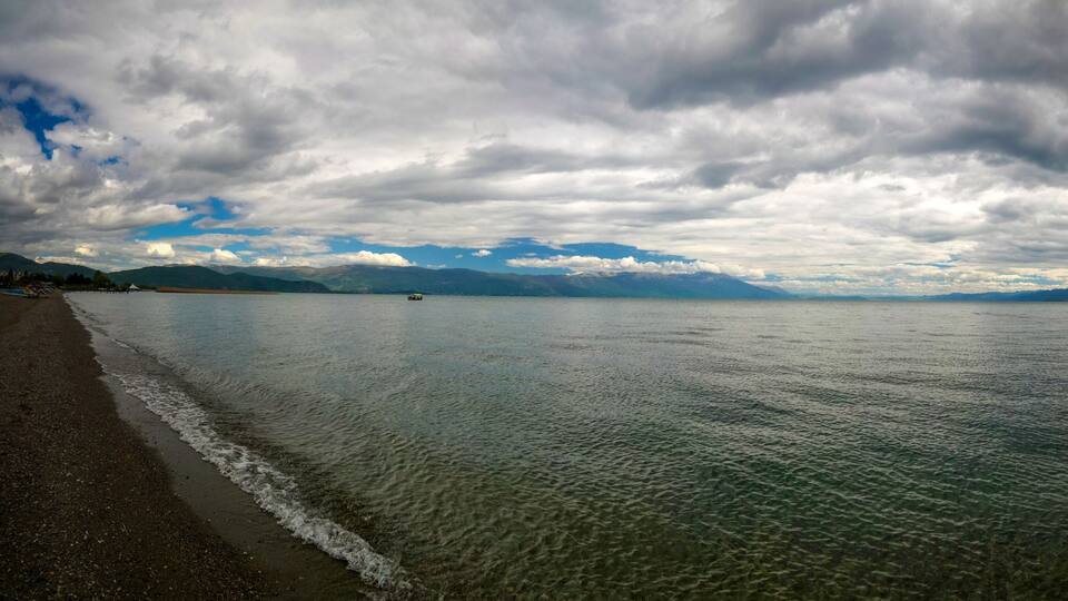A beach with calm waters, a distant mountain range, and overcast skies.
