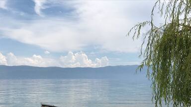A calm lake with a small boat anchored near the shore, surrounded by mountains and a willow tree.