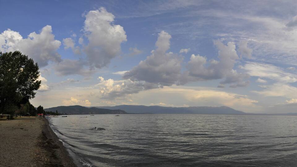 A calm lake with a distant mountain range, a partly cloudy sky, and a shoreline with sparse vegetation.