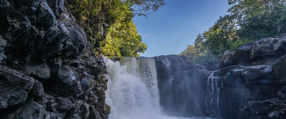 A beautiful tropical waterfall. Streams of water fall from the edge of the ledge into the river. Foam, splashes. Wet steep coastal cliffs. Green trees against a blue sky. Mauritius. GRSE waterfall