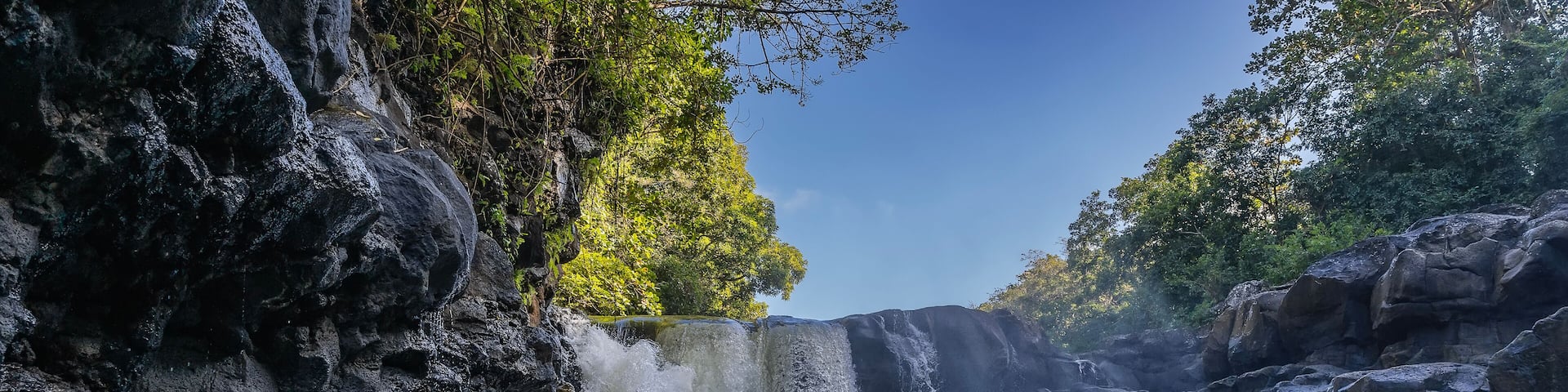 A beautiful tropical waterfall. Streams of water fall from the edge of the ledge into the river. Foam, splashes. Wet steep coastal cliffs. Green trees against a blue sky. Mauritius. GRSE waterfall