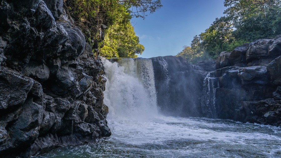 A beautiful tropical waterfall. Streams of water fall from the edge of the ledge into the river. Foam, splashes. Wet steep coastal cliffs. Green trees against a blue sky. Mauritius. GRSE waterfall