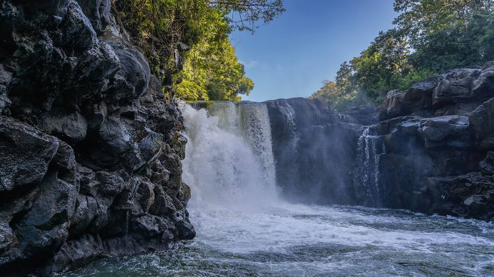 A beautiful tropical waterfall. Streams of water fall from the edge of the ledge into the river. Foam, splashes. Wet steep coastal cliffs. Green trees against a blue sky. Mauritius. GRSE waterfall