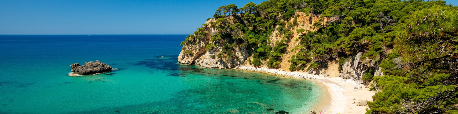 A coastal landscape with clear turquoise waters, a sandy beach, and a rocky cliff covered with greenery.