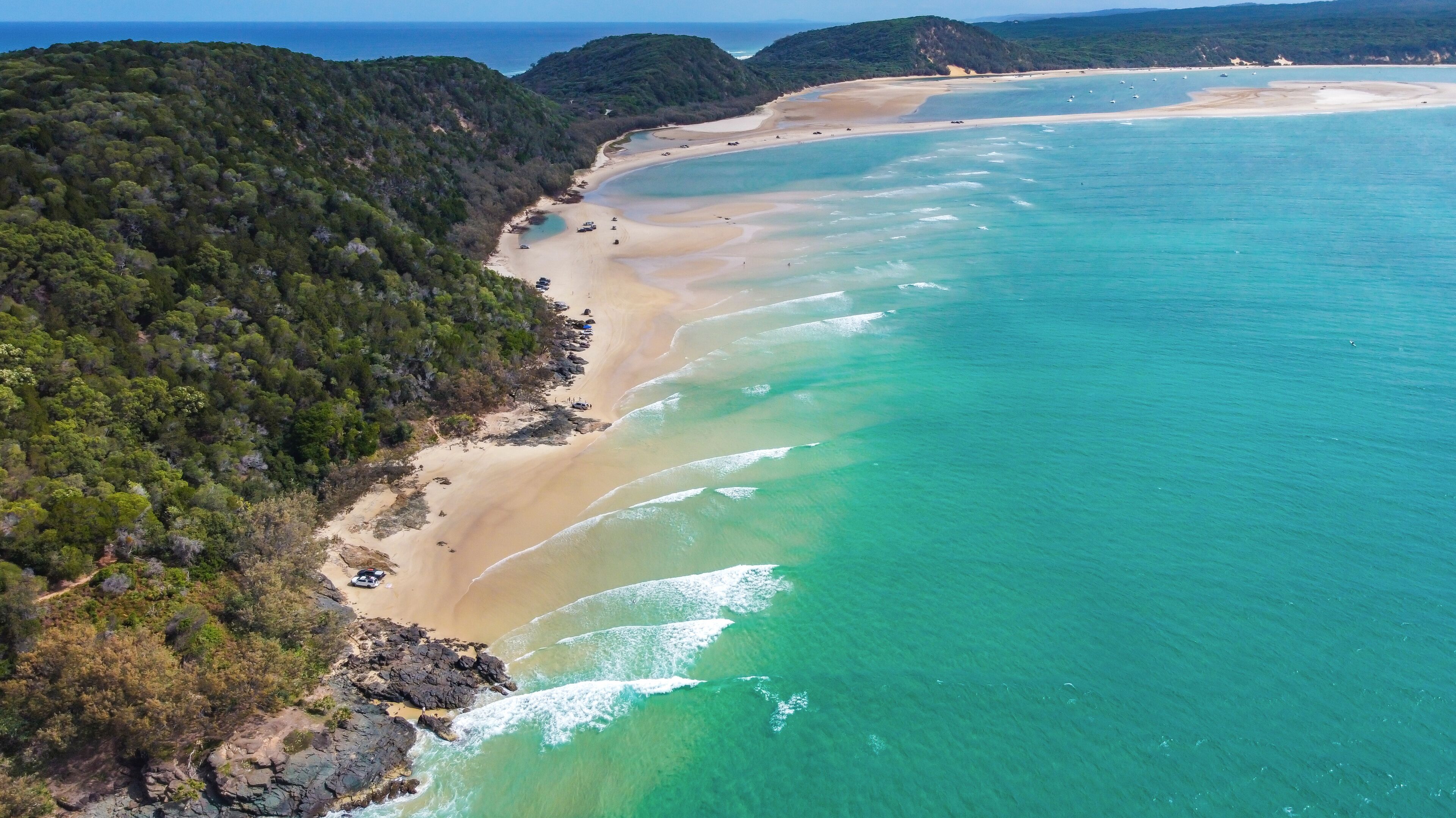Un paysage côtier caractérisé par une plage de sable, des eaux turquoise et limpides, et des collines verdoyantes et densément boisées.