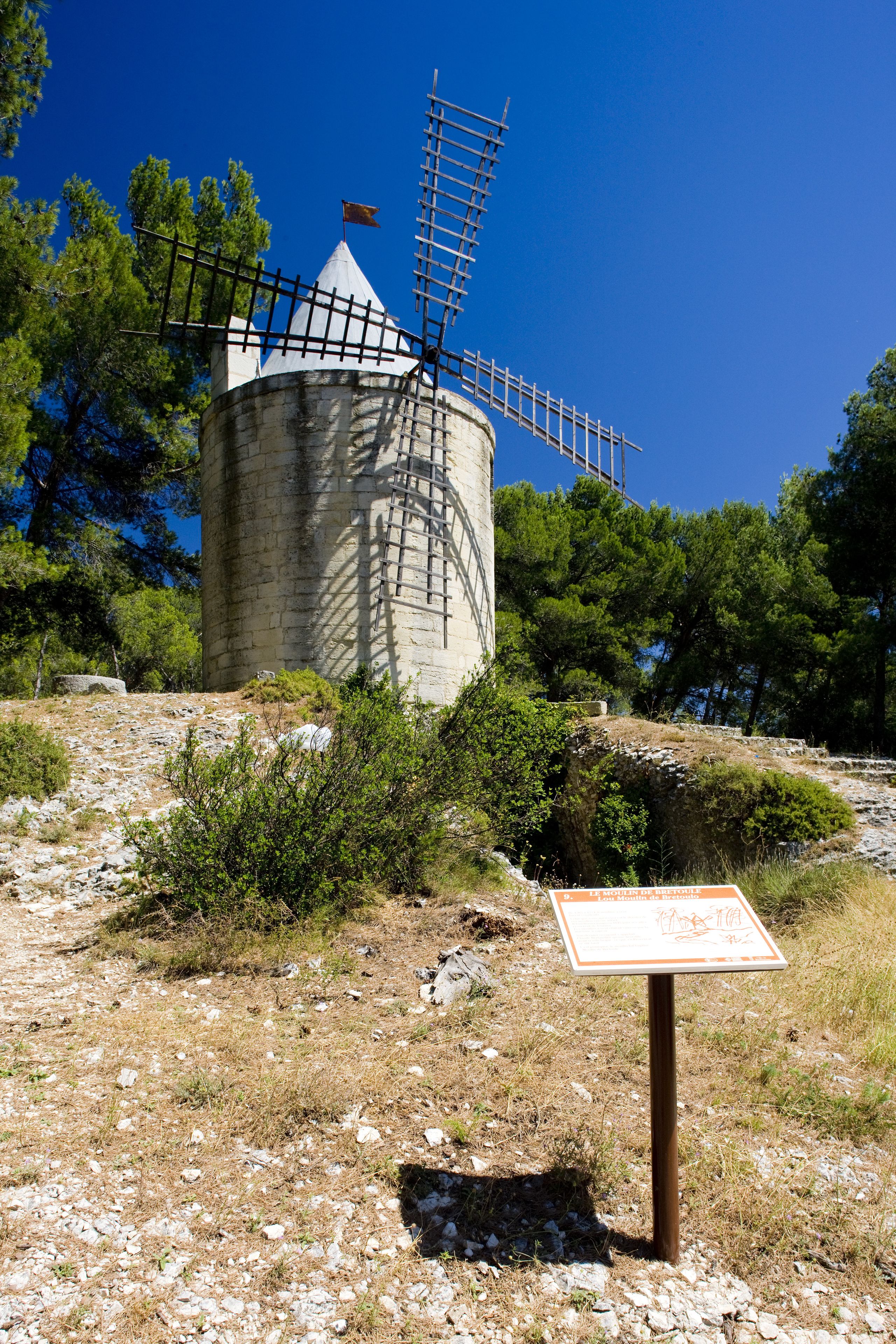 A windmill with a metal structure on top, surrounded by trees and rocky terrain.
