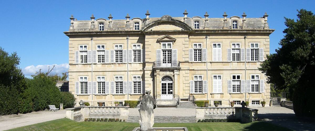 A large, historic building with symmetrical architecture, a central entrance, and a formal garden with a statue and fountains.