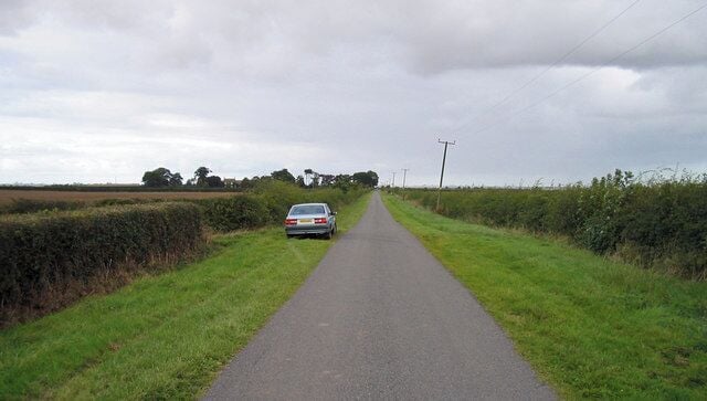 A narrow rural road flanked by fields and hedgerows under a cloudy sky.