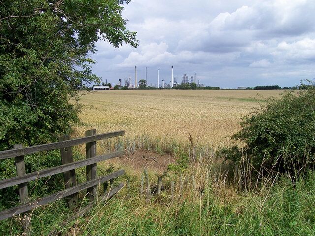 A rural landscape with a field of crops, a wooden fence, and industrial structures in the distance.
