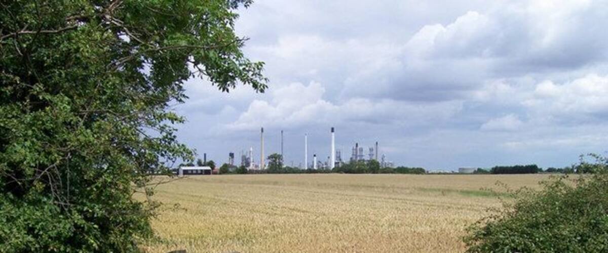A rural landscape with a field of crops, a wooden fence, and industrial structures in the distance.