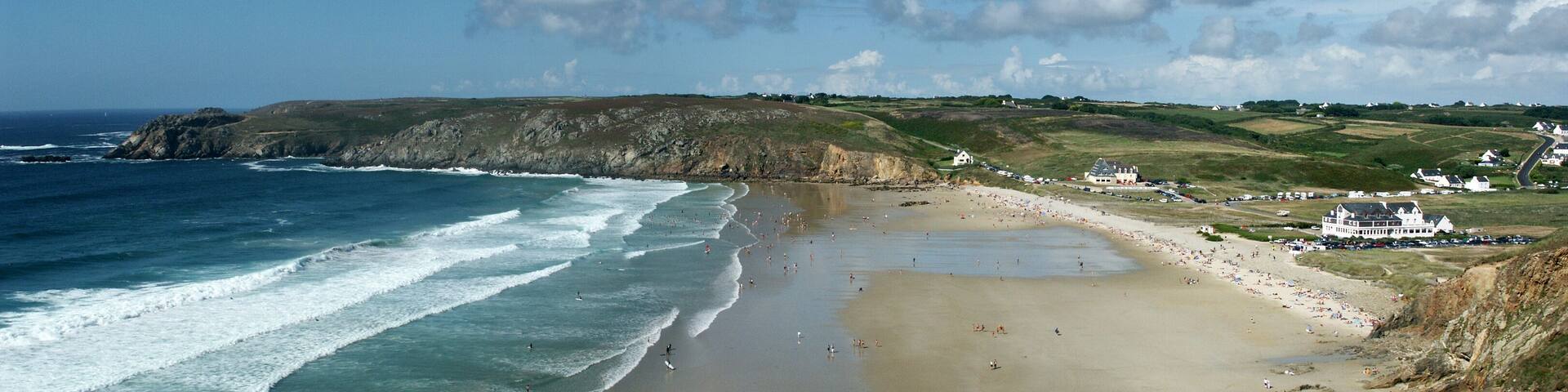 A coastal beach with a sandy shore, waves, and a grassy hillside with buildings.