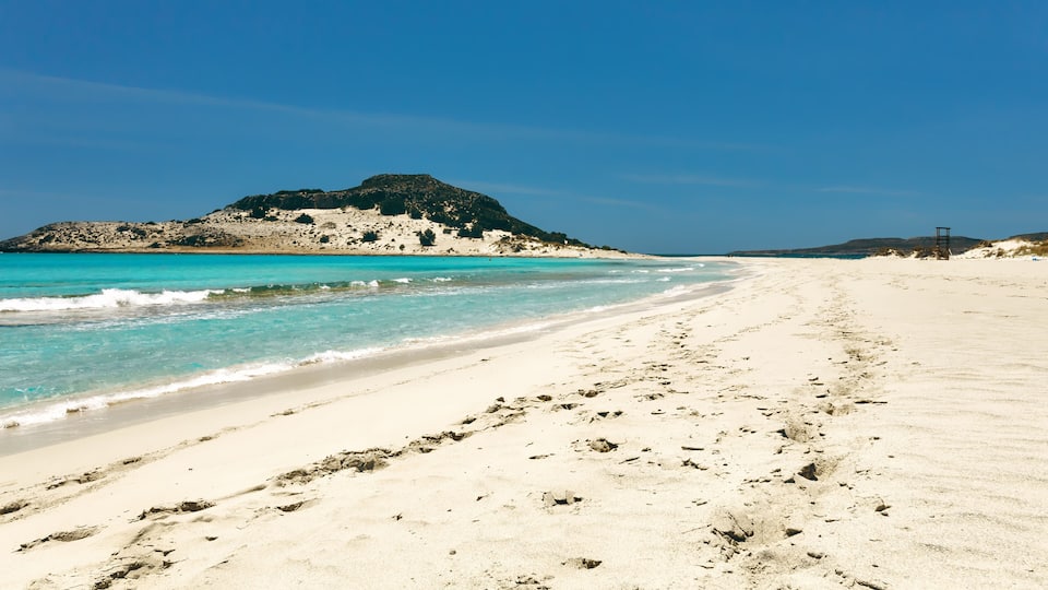 A sandy beach with clear turquoise water, footprints, and a rocky hill in the background.