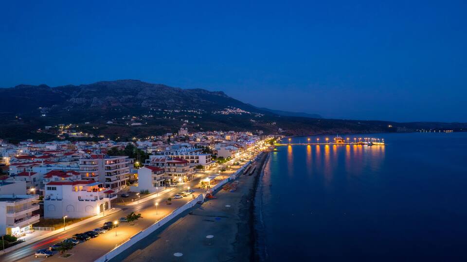 A coastal city at dusk with a long promenade, calm sea, and mountains in the background.