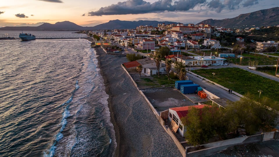 A coastal town with a pebbled beach, a boat on the water, and houses with red roofs.