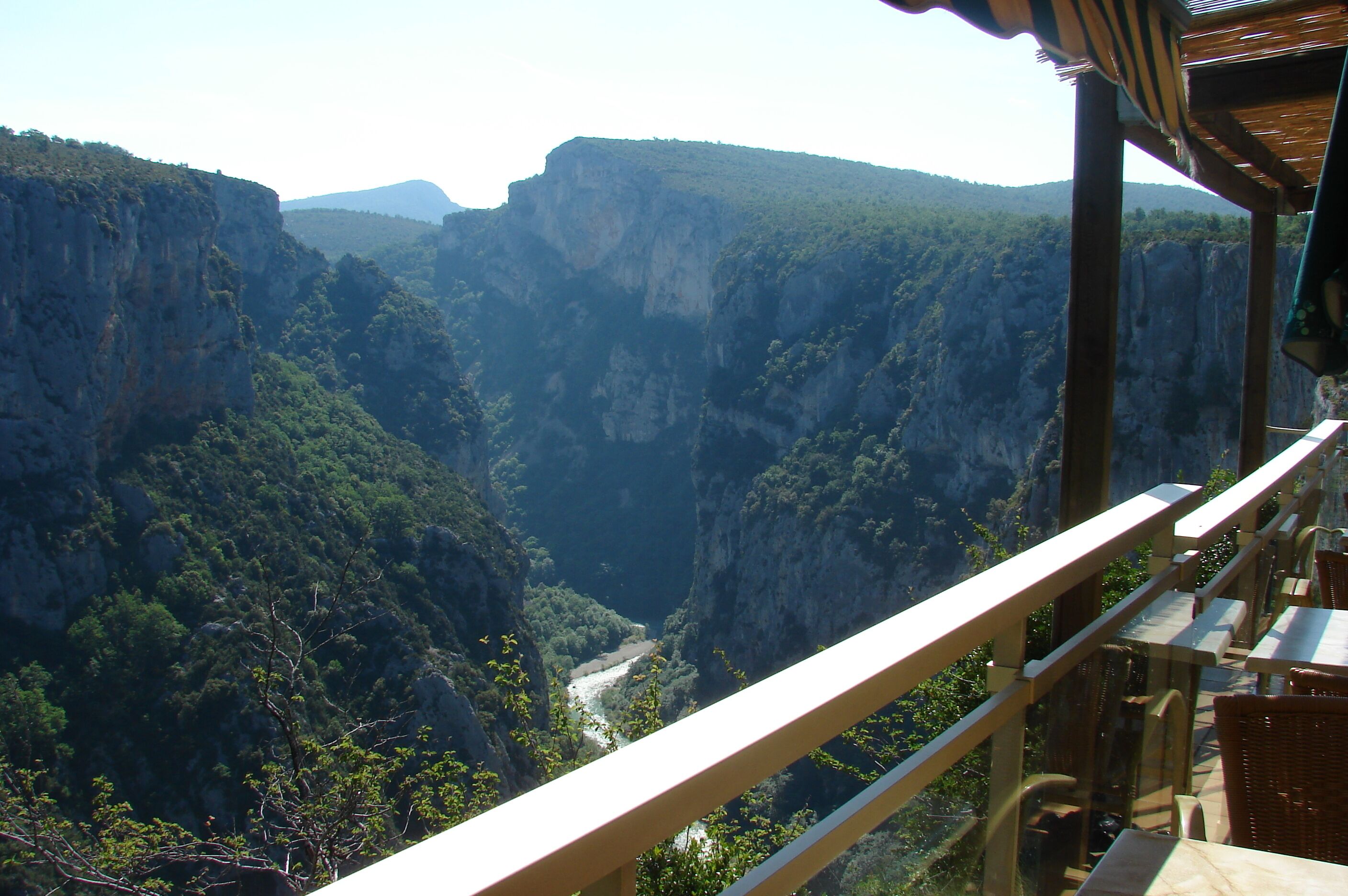 A mountainous landscape with a river flowing through a canyon, viewed from a balcony with tables and chairs.