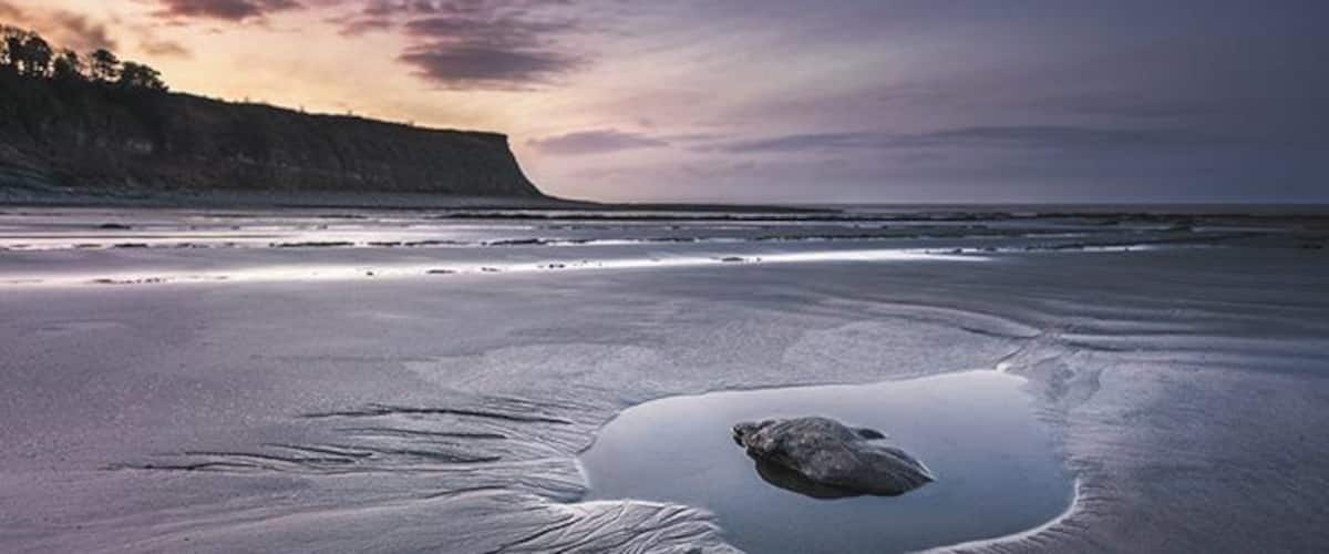 A beach with a large puddle and a rock in the center.