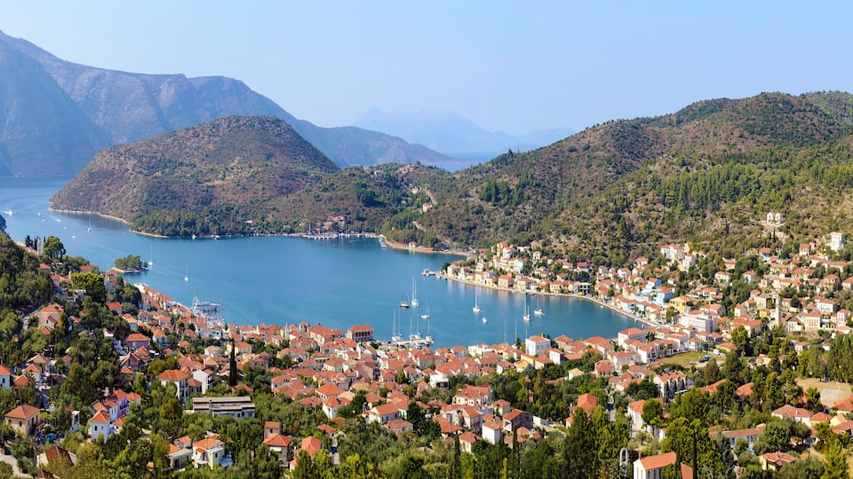 A coastal town with red-roofed houses, a bay with boats, and mountains in the background.