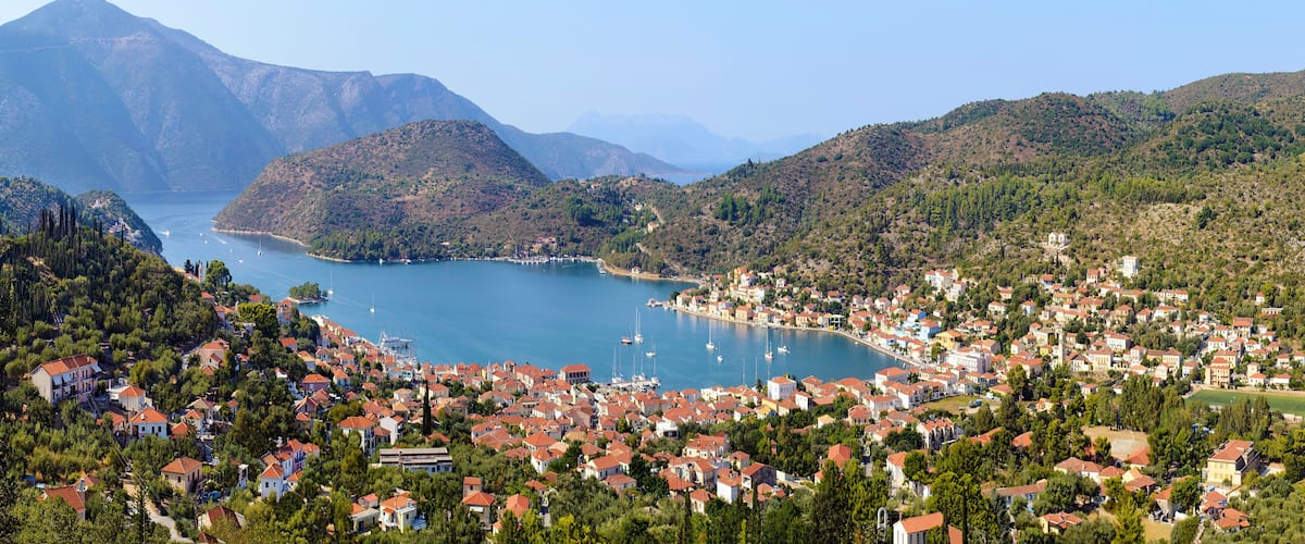 A coastal town with red-roofed houses, a bay with boats, and mountains in the background.