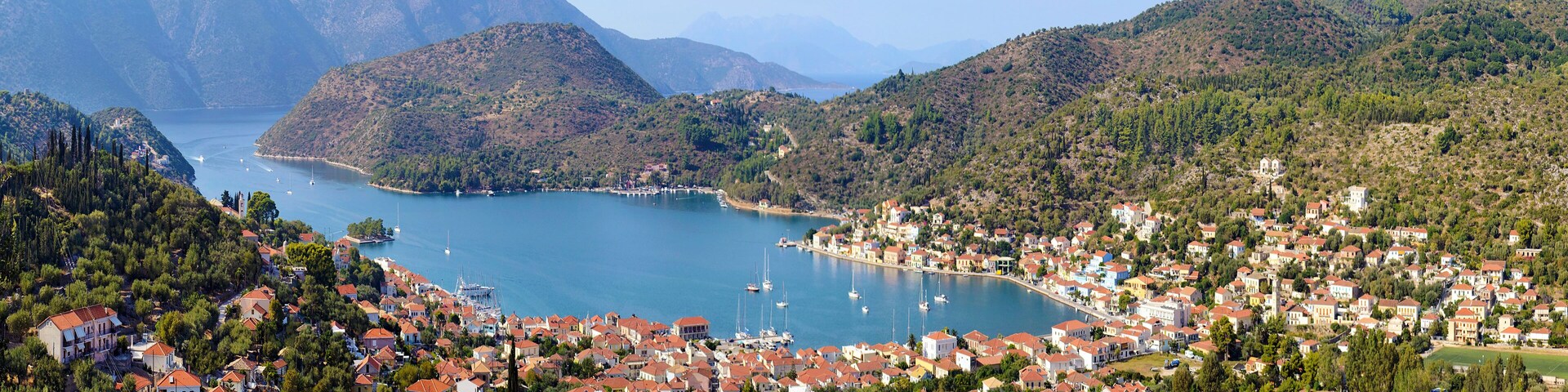 A coastal town with red-roofed houses, a bay with boats, and mountains in the background.